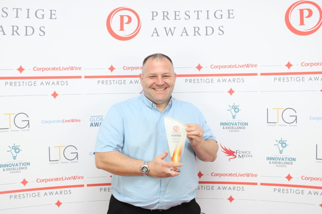 A smiling man in a light blue shirt holds a "Winner" award at the Prestige Awards event