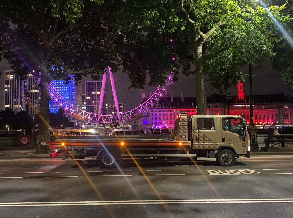 A recovery truck is parked along the road at night, illuminated by colorful lights from the London Eye